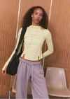 A woman stands indoors wearing a pale yellow long-sleeve cropped top with horizontal seam details, light gray drawstring sweatpants, and a black shoulder bag. The background features wood-paneled walls and a light-colored chair.