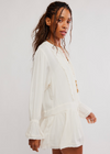 A woman wearing a white long-sleeve mini dress with ruffle trim and a tie-neck detail, photographed against a plain light gray studio background.