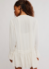 A woman viewed from behind wearing a white long-sleeve blouse with a ruffled hem and gathered back panel, photographed against a plain light background.