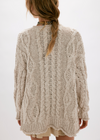 A woman viewed from behind wearing a chunky cable-knit sweater in a light oatmeal beige color, paired with a gold chain necklace and a light-colored skirt.