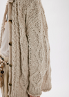 Close-up of a person wearing a chunky cream cable-knit cardigan with dark buttons over a white top and beige bottoms, against a plain light background.
