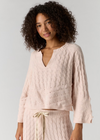 A woman with curly dark hair wearing a light blush pink cable-knit V-neck sweater and matching knit pants with a cream drawstring, photographed against a plain light gray studio background.