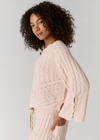 A woman with curly dark hair wearing a pale blush pink cable-knit sweater and matching knit bottoms, photographed against a light gray studio background.