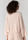 A woman shown from the back wearing a light blush pink cable-knit cardigan and matching knit pants against a plain light gray studio background.