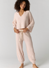 A woman modeling a light pink cable-knit lounge set, consisting of a cropped long-sleeve V-neck top and matching drawstring pants, against a plain light gray studio background.