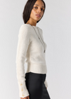 A woman wearing a cream-colored, textured cable-knit sweater with decorative buttons on the cuffs, paired with black trousers, photographed against a light gray studio background.