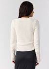 A woman shown from the back wearing a cream ribbed knit sweater and dark gray trousers, photographed against a light gray studio background.