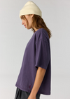 A side-profile view of a model wearing an oversized, short-sleeve purple t-shirt, a cream-colored beanie, and dark gray pants against a light gray studio background.