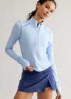 A woman modeling a light blue long-sleeve quarter-zip athletic top with contrasting light blue piping, paired with a navy wrap-style athletic skirt, against a plain light gray studio background.