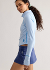 A woman modeling a light blue long-sleeve athletic top with light blue piping and a small embroidered logo, paired with a blue skirt, against a plain light gray studio background.