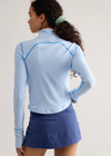 A woman shown from the back wearing a light blue long-sleeve athletic top with bright blue contrast stitching and a blue tennis skirt, against a plain light gray studio background.