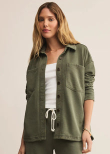  Woman wearing an olive green button-up shirt over a white top and green pants on a beige background