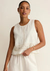 Woman wearing a white sleeveless top with lace details and white pants on a beige background