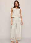 Woman wearing a light-colored sleeveless top and wide-leg pants with lace details on a plain background