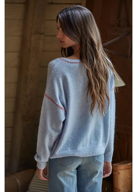 Woman wearing a light blue sweater with text, standing in a rustic indoor setting.