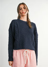 Woman wearing a navy blue cable knit sweater and pink pants with a white background