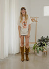 Person wearing a graphic t-shirt and brown boots in a room with a plant and rack.