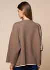 A woman shown from the back wearing a brown knit poncho-style top with contrasting light beige trim along the edges, paired with blue jeans, against a solid beige studio background.