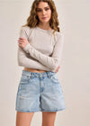 Woman wearing a beige long-sleeve top and denim shorts on a plain background