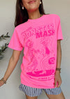A woman wearing a bright pink graphic t-shirt with 'MONSTER MASH' and dancing cartoon characters printed on the front, paired with black and white striped shorts, standing against a textured off-white wall.