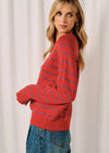 A woman wearing a red knit sweater with thin blue horizontal stripes, paired with blue jeans, standing against a white curtain background.