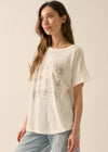 A woman wearing an oversized off-white t-shirt with a black line art graphic of cartoon faces, paired with light blue jeans, set against a plain beige background.