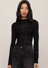 Woman wearing a black long-sleeve top with intricate patterns and high-waisted black jeans on a beige background