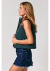 A woman shown in profile wearing a sleeveless dark teal-green puffer vest with brown faux-leather trim over a dark top and a denim skirt, against a light gray studio background.