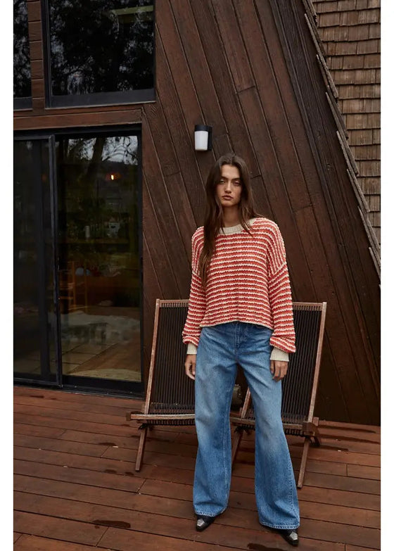 Woman in a striped sweater and blue jeans standing on a wooden deck with a wooden building in the background.