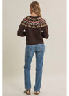 A woman viewed from behind wearing a dark brown knit sweater with a colorful geometric pattern across the shoulders and blue jeans, standing against a plain beige studio background.