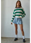 Person wearing a green and white striped sweater with denim shorts against a plain background