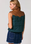 A woman shown from the back wearing a sleeveless quilted vest with a dark teal body and brown shoulder yoke, paired with a denim skirt, against a light gray studio background.
