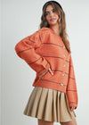 Woman wearing a rust-colored sweater with black stripes and a beige pleated skirt on a white background