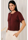 A woman modeling a short-sleeve maroon knit cardigan with a button front, paired with white pants, set against a soft white curtain background.