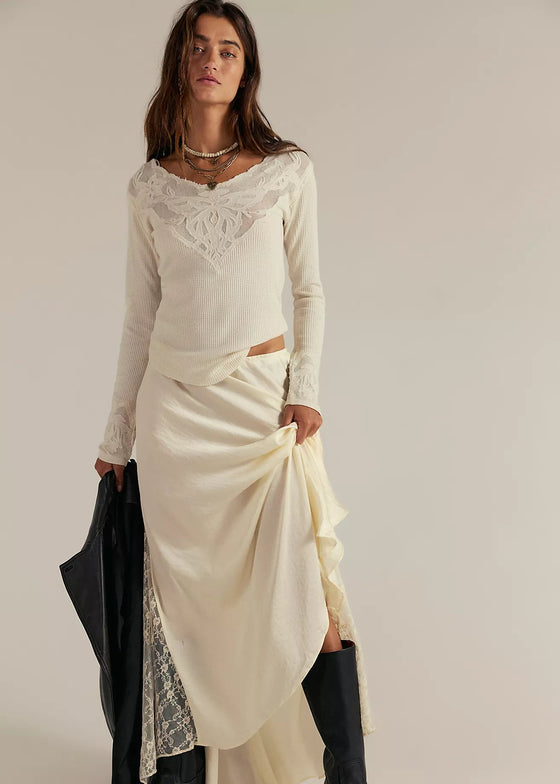 Woman wearing a long-sleeve white top and cream skirt with black boots on a plain background