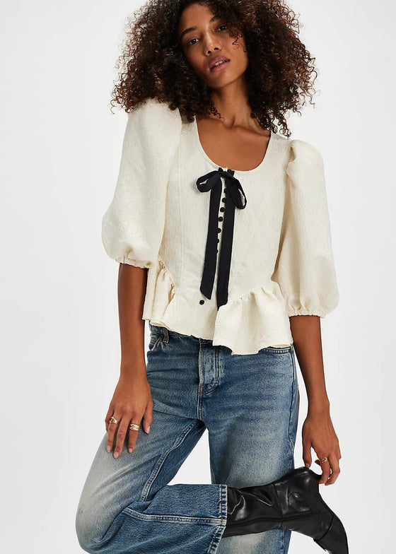 Woman wearing a white blouse with black ribbon details, blue jeans, and black boots on a white background