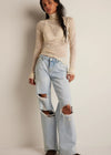 Woman wearing a beige turtleneck and light blue ripped jeans against a plain background