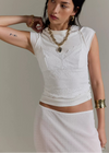 Woman wearing a white lace top and skirt with gold jewelry against a neutral background