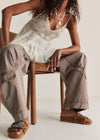 Woman sitting on a chair wearing a light-colored sleeveless top and beige pants, with a neutral background.