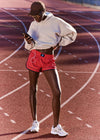 Person on a running track wearing a white sweatshirt, red shorts, and white sneakers, using a phone.