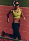 Woman running on a track wearing a green sports bra and black leggings.