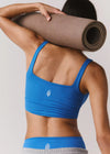 Person wearing a blue sports bra and gray leggings holding a yoga mat on a plain background