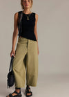 Woman wearing a black sleeveless top and beige pants, holding a black bag on a plain background
