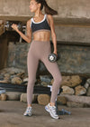 A woman in a gym setting wearing a black and taupe sports bra and matching high-waisted taupe leggings, holding dumbbells while standing on concrete.