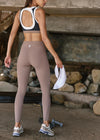 A woman from behind wearing high-waisted taupe leggings and a black, white, and beige sports bra, standing outdoors near a rustic stone structure and holding a white towel.