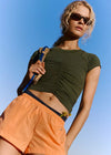 A woman wearing a dark olive green ruched short-sleeve crop top and peach-orange shorts, holding a blue and beige shoulder bag against a clear blue sky.