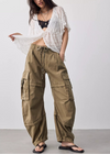 A woman modeling loose, wide-leg olive cargo pants with a drawstring waist, styled with a white sheer open-front top, black bralette, and black sandals against a plain light gray studio background.