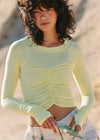 A woman wearing a pale yellow-green long-sleeve ruched crop top and white pants, standing outdoors against a sandy beige background.
