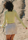 A woman seen from behind on a sandy beach, wearing a pale yellow long-sleeve crop top and white shorts.