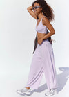 A woman modeling a light purple/lavender activewear set consisting of a wrap-style sports bra and matching wide-leg sweatpants, styled with white sneakers and sunglasses against a bright white background.